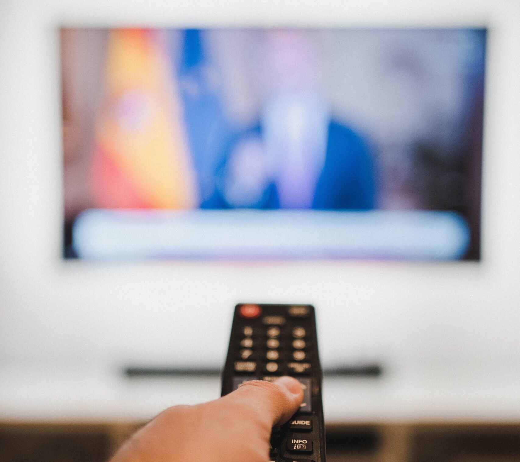 Close-up of a hand holding a remote control towards a TV screen indoors.